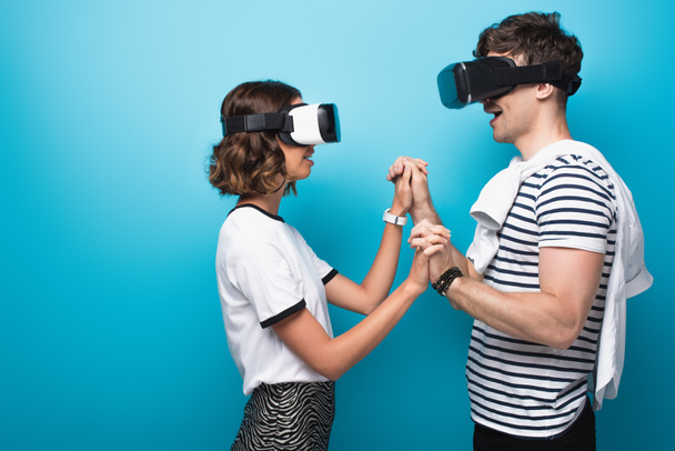 young man and woman holding hands while using virtual reality headsets on blue background - Valokuva, kuva