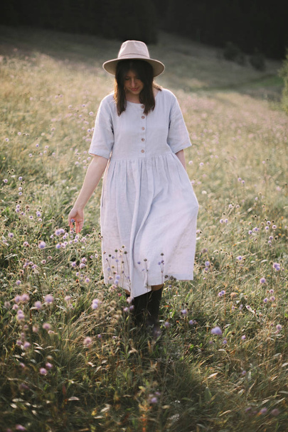 日当たりの良いメアで野生の花の中を歩く素朴なドレスのスタイリッシュな女の子 - 写真・画像