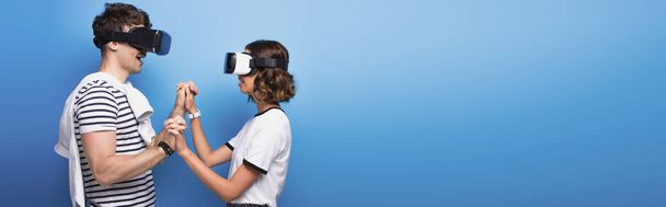 panoramic shot of young man and woman holding hands while using virtual reality headsets on blue background - Foto, afbeelding