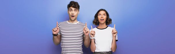 panoramic shot of cheerful man and woman looking up and pointing with fingers on blue background - Foto, imagen