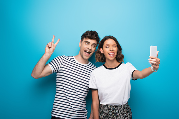 cheerful girl sticking tongue out while taking selfie with cheerful man showing victory sign on blue background - Foto, immagini