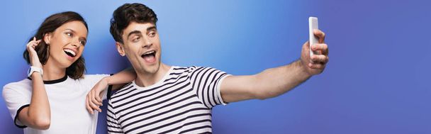panoramic shot of cheerful man taking selfie with attractive, smiling girl on blue background - Foto, afbeelding