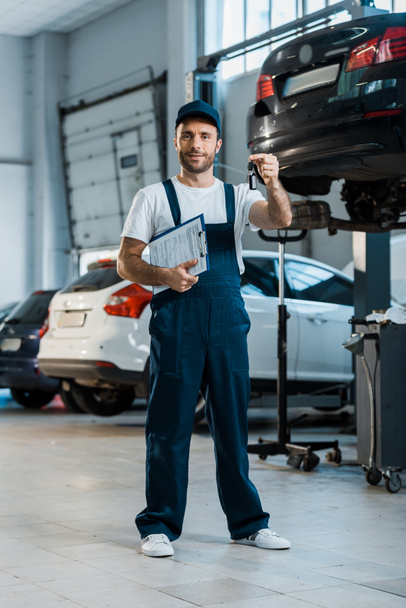 happy car mechanic looking at camera and holding clipboard and key near cars  - Fotoğraf, Görsel