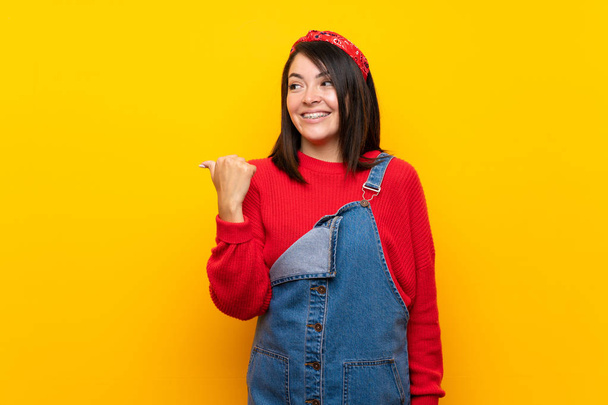 Young Mexican woman with overalls over yellow wall pointing to the side to present a product - Фото, изображение