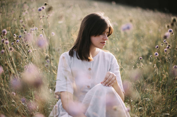 Chica con estilo en vestido rústico sentado entre flores silvestres y hierbas
 - Foto, Imagen