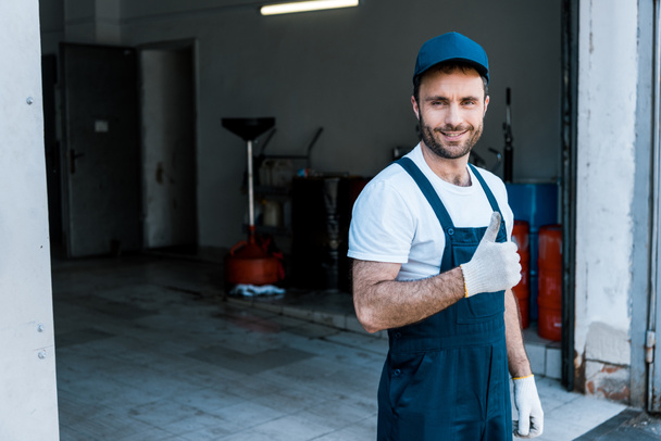 handsome bearded car mechanic in cap standing and showing thumb up  - Fotografie, Obrázek