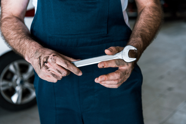 Ausgeschnittene Ansicht von Automechaniker mit Schlamm auf Händen, der einen Handschlüssel in der Autowerkstatt hält  - Foto, Bild