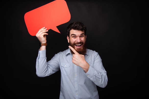 Photo of handsome bearded man holding a empty bubble speech, looking at camera while pointing at red blank - Photo, image