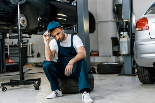 upset bearded man touching cap and sitting on car tire  - Fotografie, Obrázek