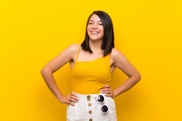 Young Mexican woman over isolated yellow background posing with arms at hip and smiling - Fotoğraf, Görsel