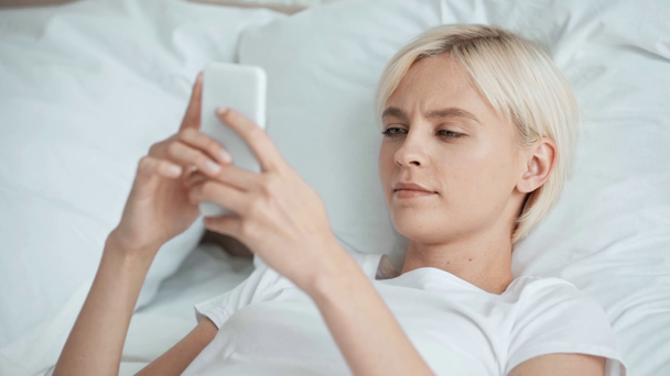 piękna dziewczyna w piżamie za pomocą smartfona i uśmiechając się w łóżku  - Materiał filmowy, wideo