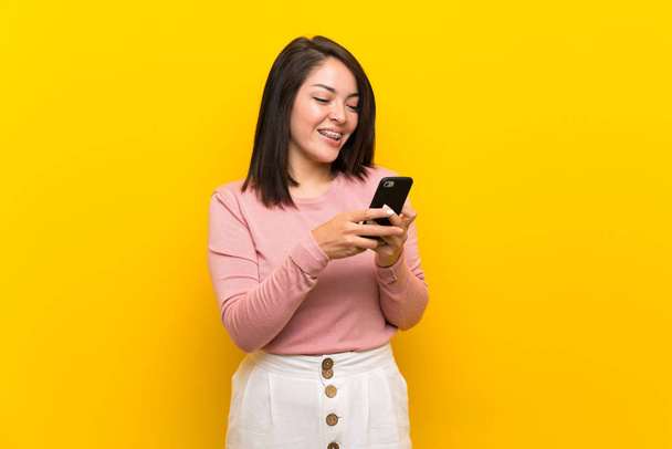 Joven mexicana sobre aislado fondo amarillo enviando un mensaje con el móvil
 - Foto, Imagen