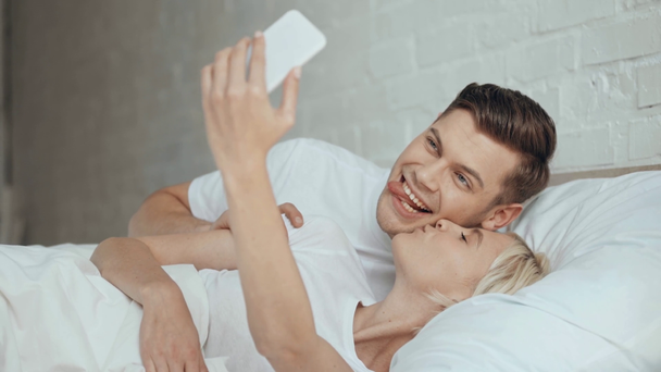 beautiful couple sticking tongue out, smiling, kissing and taking selfie on smartphone in bed - Footage, Video