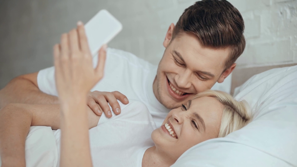 Schönes Paar streckt Zunge heraus, lächelt, küsst und macht Selfie auf Smartphone im Bett - Filmmaterial, Video