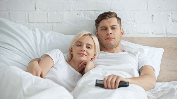 ζευγάρι κοιτάζοντας την κάμερα, άνθρωπος αγκάλιασμα γυναίκα και αλλαγή καναλιών με τηλεχειριστήριο στο κρεβάτι - Πλάνα, βίντεο