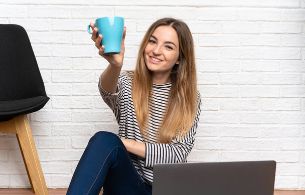 Mujer joven sentada en el suelo con su portátil sosteniendo una taza de café
 - Foto, imagen