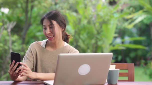 Aasialainen nainen käyttää matkapuhelimen selfie post sosiaalisessa mediassa, nainen rentoutua onnellinen näyttää ostoskassit istuu pöydällä puutarhassa aamulla. Elämäntapa naiset rentoutua kotona käsite. - Materiaali, video