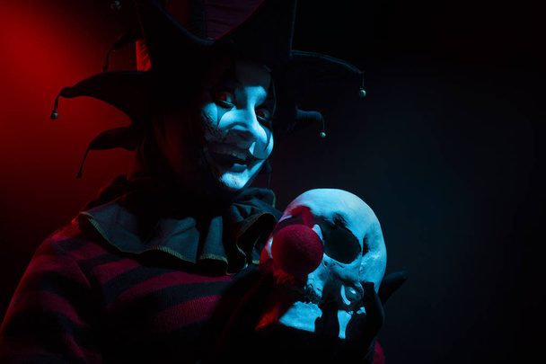 Ανατριχιαστικός γελωτοποιός που κρατά ένα ανθρώπινο κρανίο με κόκκινη μύτη κλόουν. - Φωτογραφία, εικόνα