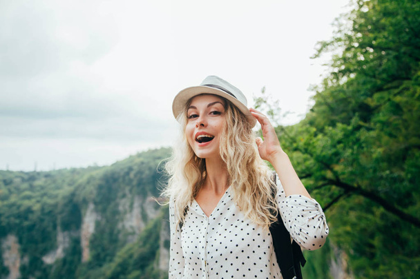 cheerful lady with blond flowing hair in a big white polka-dot shirt holding a hat with her hand, a happy young woman smiles broadly, a natural make-up, a cute gentle image of a girl tourist - Zdjęcie, obraz