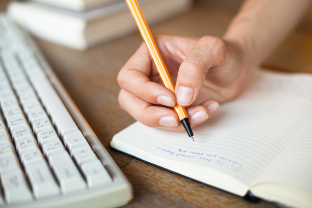 χέρια γράφει ένα στυλό σε ένα σημειωματάριο, πληκτρολόγιο του υπολογιστή στο παρασκήνιο - Φωτογραφία, εικόνα