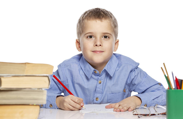Aranyos fiú iskolás korú csinál házi feladatot az asztalnál. Érdekes megtanulni. Fehér alapon izolált. - Fotó, kép