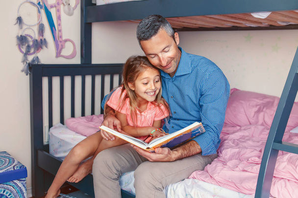 Family two people sitting on bed in bedroom reading book. Father and daughters girl at home spending time together. Parent talking communicate to child. Real people authentic lifestyle. - Photo, Image