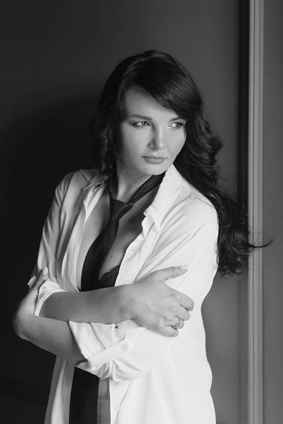 μονόχρωμος πορτρέτο του κομψό, όμορφη μελαχρινή γυναίκα με μακριά μαλλιά σε μαύρο σουτιέν και γραβάτα σε λευκό πουκάμισο και λευκές πένες - Φωτογραφία, εικόνα