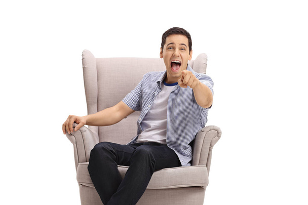 Young guy sitting in an armchair pointing at the camera and laughing out loud isolated on white background - Foto, imagen
