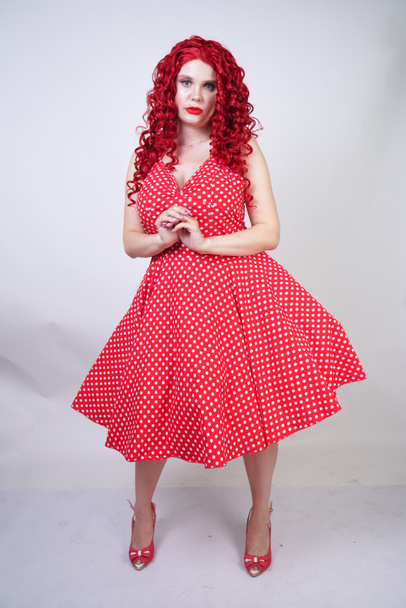 plus size red hair curly woman with curvy body wearing retro polka dot pretty dress and standing on white studio background alone. chubby fun fashion girl in vintage clothes posing. - Fotoğraf, Görsel