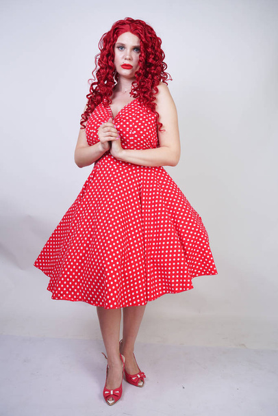 plus size red hair curly woman with curvy body wearing retro polka dot pretty dress and standing on white studio background alone. chubby fun fashion girl in vintage clothes posing. - Photo, image
