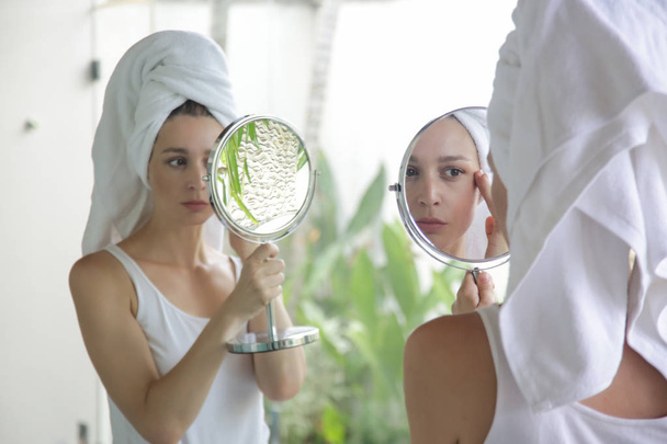 Πορτραίτο μιας όμορφης νεαρής γυναίκας με μια πετσέτα στο κεφάλι που στέκεται στο μπάνιο και εξετάζοντας το πρόσωπό της στον καθρέφτη, τη φροντίδα του δέρματος και την έννοια του αντι-ηλικίας - Φωτογραφία, εικόνα