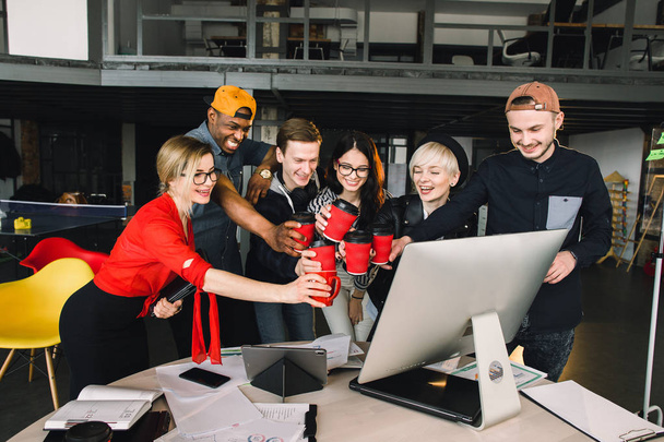 Group of international students or workers in loft office clinking cups of coffee, six colleagues of modern work co-working space talking and smiling while standing at the desk with laptops - 写真・画像