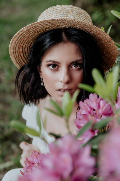 Close up portrait of a beautiful girl in a white vintage dress and straw hat standing near pink flowers - Foto, Imagem