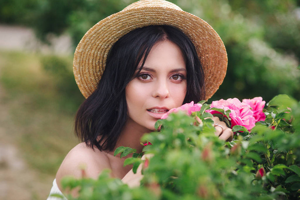 ストロー帽子のロマンチックなクローズアップポートレートo魅力的なブルネットの女の子は、バラの茂みで花の匂いを嗅ぎます - 写真・画像