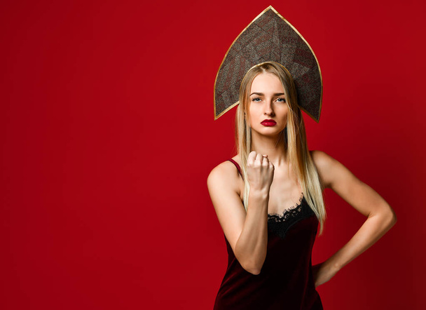 Angry beautiful young woman in kokoshnik hat shows fist at camera, threats someone dressed in velvet dress, isolated over red background, - Foto, immagini