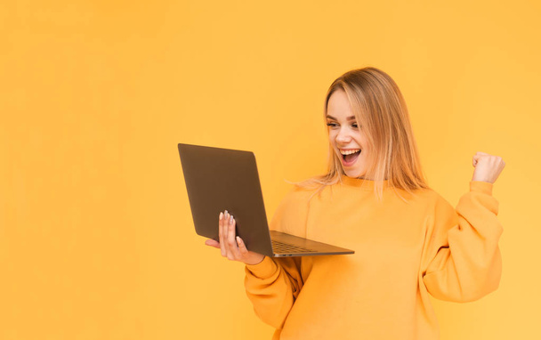 Portrait of a teenage girl standing on a yellow background with a laptop in her hand, rejoices and looks at the screen. Happy blond girl uses a laptop isolated. Copy space - 写真・画像