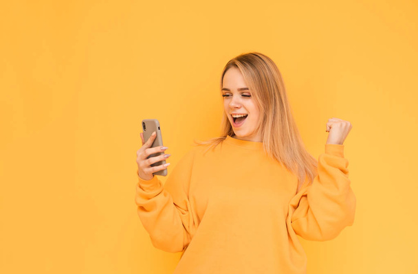 笑顔のブロンドの女の子は明るい服でスマートフォンを使用し、黄色の背景に喜びます。スマートフォンの画面を見て幸せな女の子の肖像画と勝利に喜び、孤立. - 写真・画像