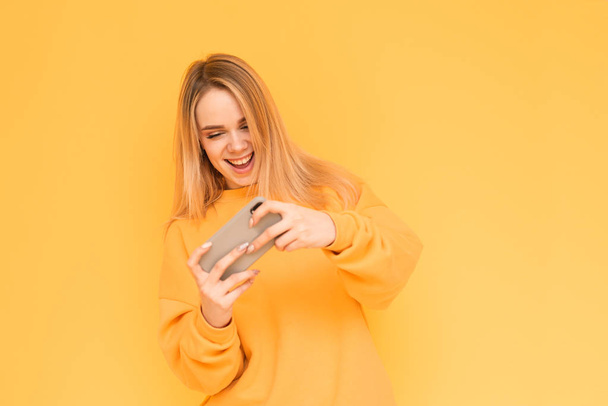 Portrét mladé dívky v oranžových šatech hrající mobilní hry na smartphone s úsměvem, dívajíce se na obrazovku telefonu na žlutém pozadí. Mobilní hraní. Holka je Gamer. Izolované. - Fotografie, Obrázek