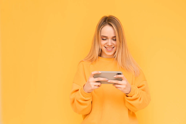 Portrait of a smiling girl in bright orange clothes is with a smartphone in her hands on a yellow background, looking at the screen and smiling. Happy girl podilot plays mobile games on a smartphone. - 写真・画像