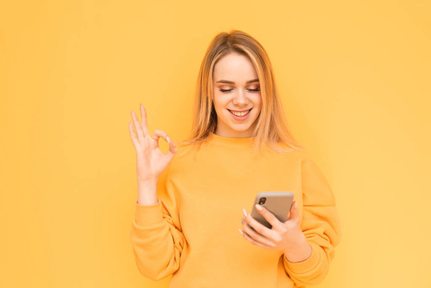 Happy girl in bright clothes uses a smartphone on a yellow background and shows an OK gesture with her fingers. Smiling blonde uses the internet on a smartphone, isolated on yellow. Copy space - Foto, immagini
