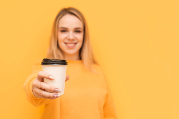 Happy girl holds a paper glass of coffee in her hand and shows her in the camera on a yellow background, smiling and looking. Focus on a cup of coffee in the hands of a young girl underground. - Foto, afbeelding