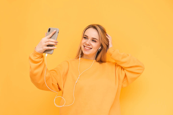 笑顔の美少女はセフィを作り、明るいセーターを着て黄色の背景にヘッドフォンで音楽を聴きます。ブロンドモデルは、スマートフォン、耳のヘッドフォンで彼女の写真を作ります - 写真・画像