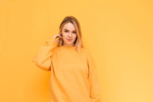 Modelo atraente em uma camisola laranja posa, sorri e olha para a câmera, isolada em um fundo amarelo. Senhora positiva fica no fundo de uma parede amarela, sorrisos bonitos
. - Foto, Imagem