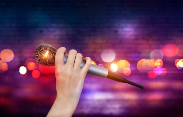Ένα θηλυκό χέρι κρατά ένα μικρόφωνο με τα πολύχρωμα φώτα της σκηνής του καραόκε κλαμπ. Φωτεινό πολύχρωμο φόντο με θολά φώτα νέον - Φωτογραφία, εικόνα