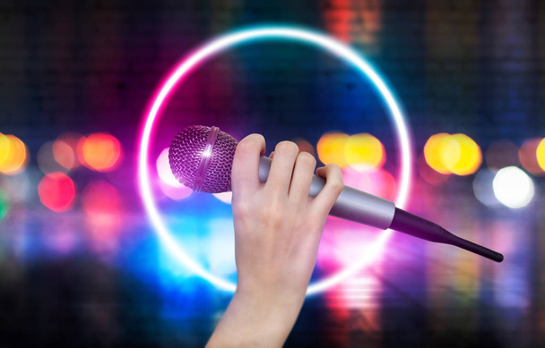 Жіноча рука тримає один мікрофон проти барвистих вогнів сцени караоке-клубу. Яскравий барвистий фон з розмитими неоновими вогнями
 - Фото, зображення