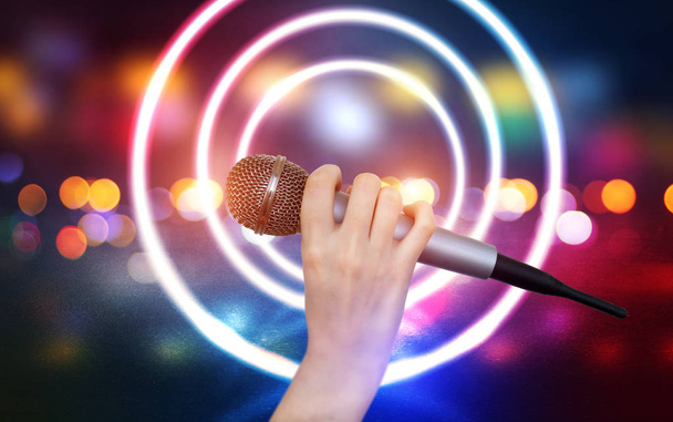 Egy női kéz egy mikrofonnal a karaoke klubélet színes fényei ellen. Élénk színes háttér homályos neonfények - Fotó, kép