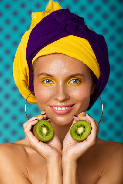 Γαλανομάτα νεαρή χαμογελαστή γυναίκα με πολύχρωμο περιτύλιγμα στο κεφάλι και καλοκαιρινό μακιγιάζ που κρατάει δύο μισά ακτινίδια φρούτων. Κοντινό πορτραίτο ομορφιάς. - Φωτογραφία, εικόνα