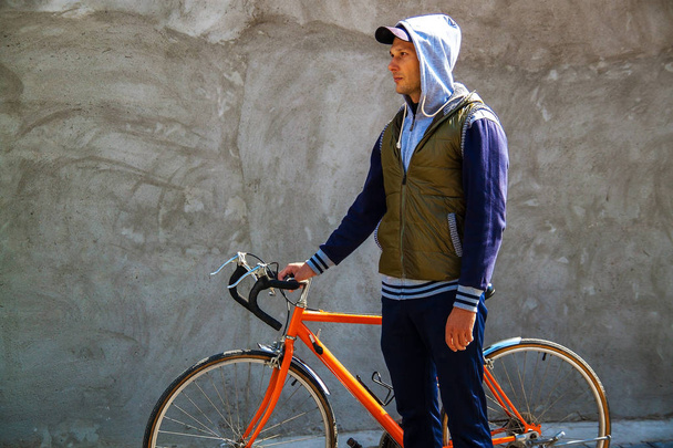 Πορτραίτο ενός χαμογελαστή νεαρού άντρα που στέκεται με το ποδήλατό του μπροστά από ένα γκρίζο τοίχο στην πόλη το φθινόπωρο με χώρο αντιγραφής. - Φωτογραφία, εικόνα