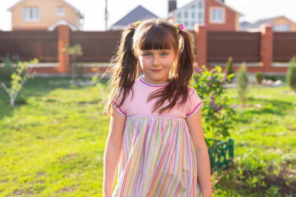 Niña pequeña en vestido rosa en el patio mira a la cámara y sonríe
 - Foto, Imagen