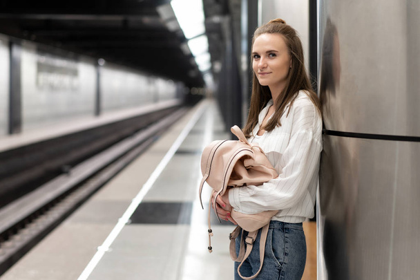 young european girl in jeans and shirt with backpack is waiting for the train at the metro station. Background in motion blur to convey a vibrant atmosphere. This is a snapshot idea. - Photo, Image